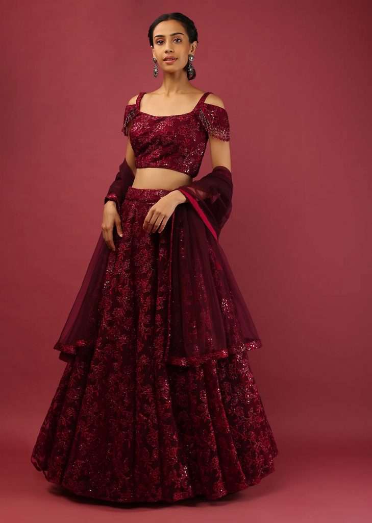 A woman poses for a photo wearing a maroon lehenga choli in an embroidered net with beaded fringe on cold shoulder sleeves
