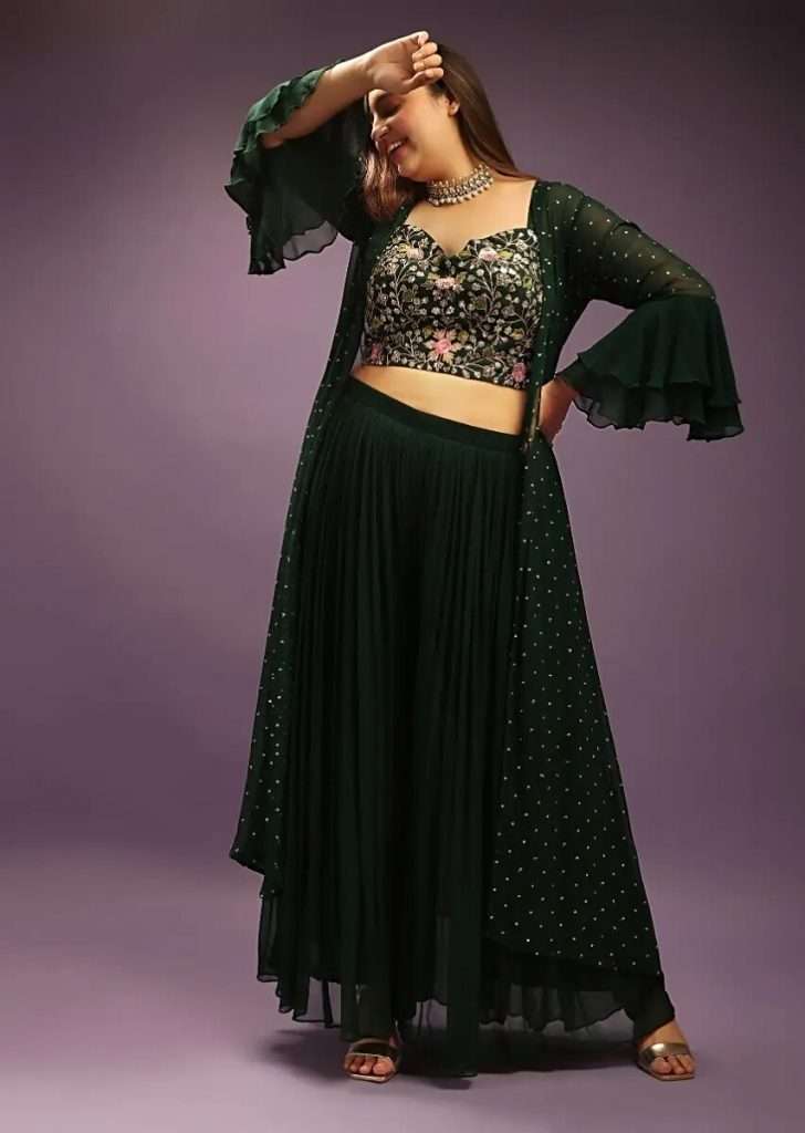 A woman in a green palazzo suit with a matching jacket and embroidered floral net poses for a photograph.
