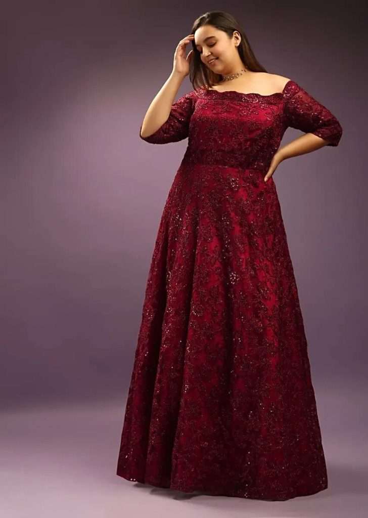 A woman in a ruby maroon off-shoulder gown adorned with embroidered thread and sequins poses for the photo