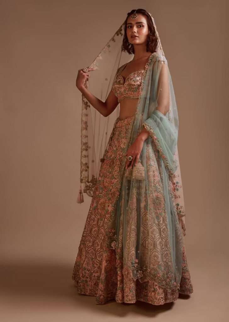 A woman wearing a powder pink lehenga in heavily hand-embroidered raw silk in floral and Moroccan motifs with 3D flower detailing poses for a photo