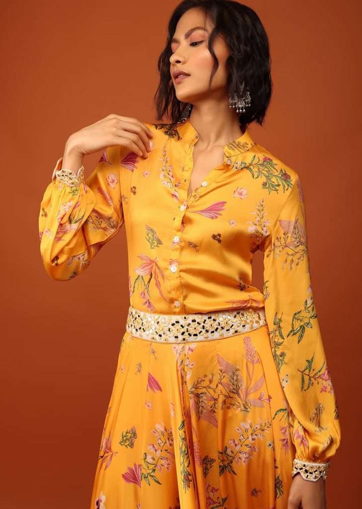 A woman in a saffron yellow floral print shirt top and mirror embroidered lehenga poses for a photo