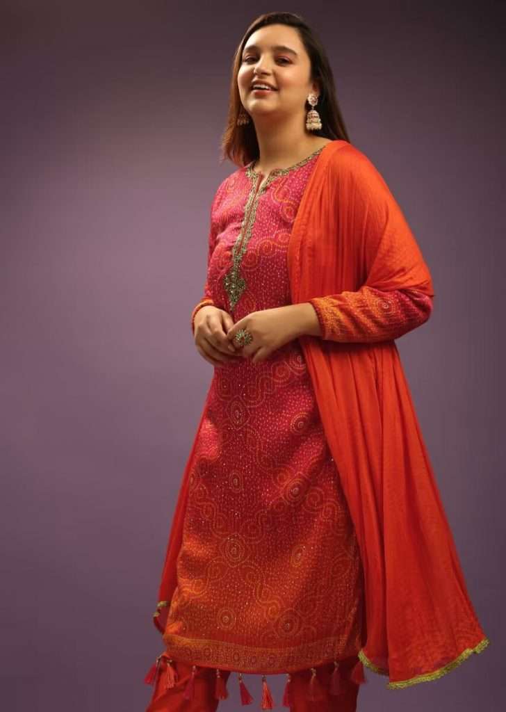 A woman is photographed in a hot pink and orange straight cut chiffon suit with bandhani design all over and tassels at the hem