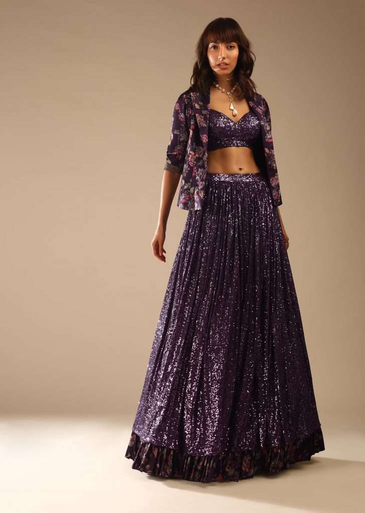 A woman is photographed wearing a floral printed blazer jacket and sequin embellished jeweled purple lehenga choli with printed frill at the hemline of the lehenga